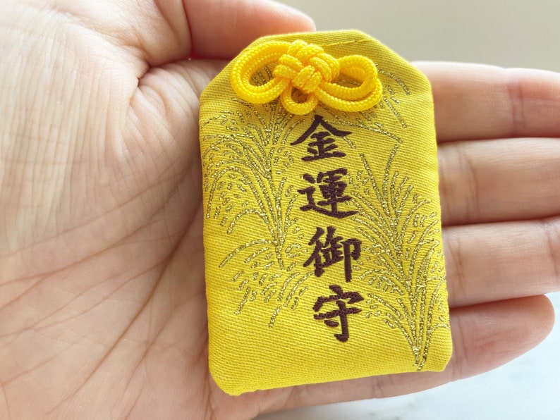 May include: A yellow Japanese omamori charm with gold accents and kanji characters. The charm has a decorative knot at the top and a gold embroidered design. The text on the charm is in dark brown.