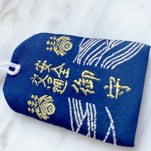 May include: A navy blue Japanese omamori charm with gold embroidered text and floral designs. The charm has a white string attached for hanging. The design includes white embroidered wave patterns.