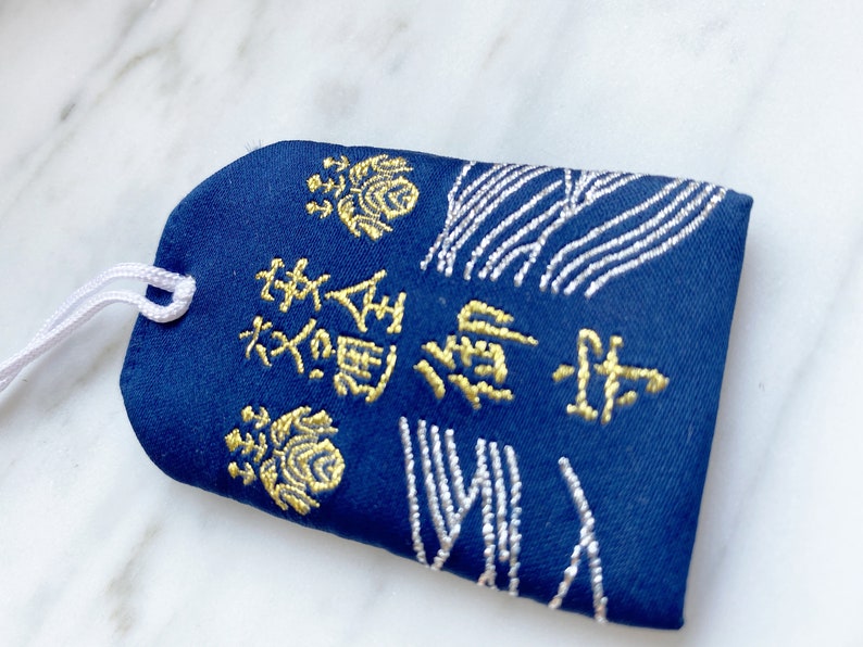 May include: A navy blue Japanese omamori charm with gold embroidered text and floral designs. The charm has a white string attached for hanging. The design includes white embroidered wave patterns.
