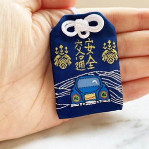 May include: Navy blue Japanese omamori charm with white accents. The charm features gold embroidery and the Japanese characters for traffic safety. A blue car is embroidered on the bottom. A white cord and bow are at the top.