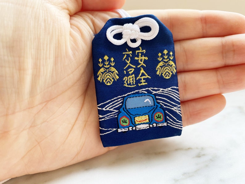 May include: Navy blue Japanese omamori charm with white accents. The charm features gold embroidery and the Japanese characters for traffic safety. A blue car is embroidered on the bottom. A white cord and bow are at the top.