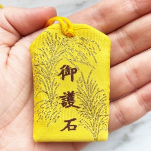 May include: Yellow Japanese omamori charm with gold accents and Japanese characters. The charm has a gold loop for hanging and features a decorative design. The text on the charm is in a dark brown color.