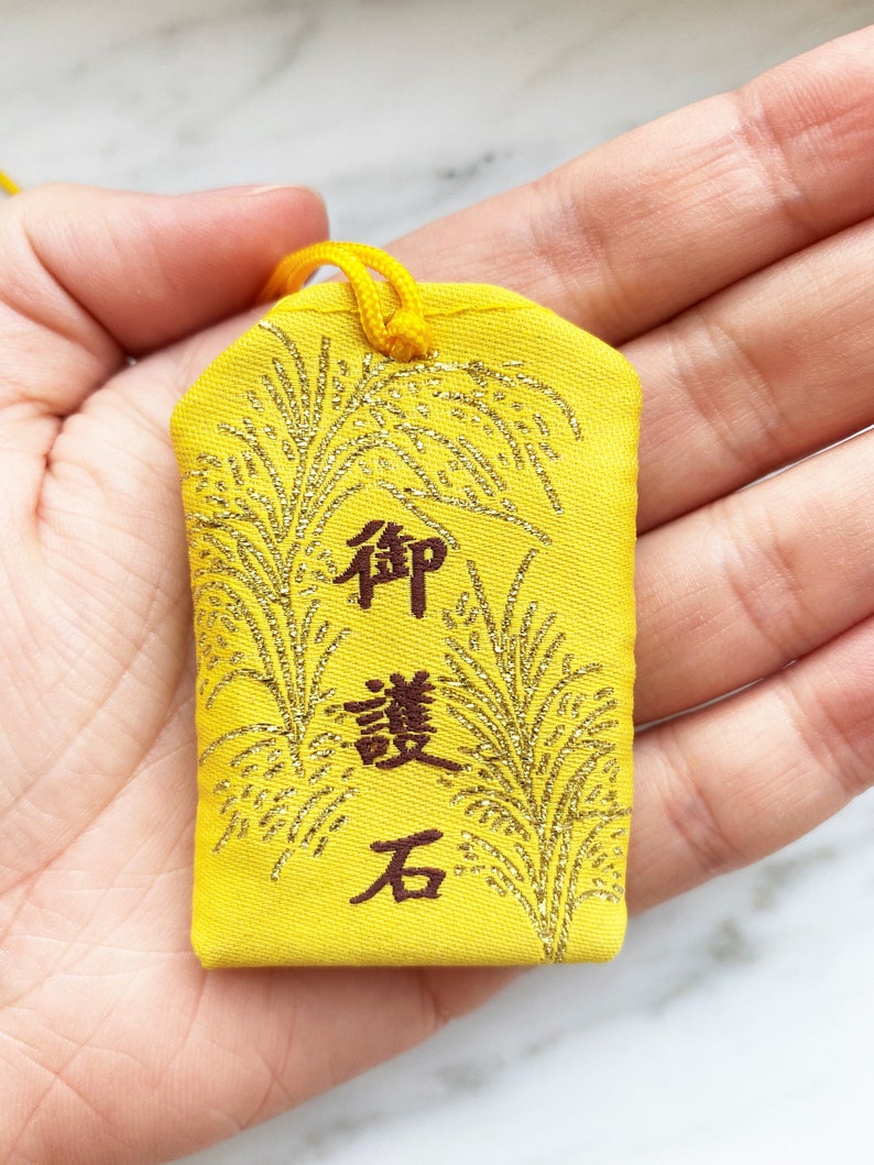 May include: Yellow Japanese omamori charm with gold accents and Japanese characters. The charm has a gold loop for hanging and features a decorative design. The text on the charm is in a dark brown color.