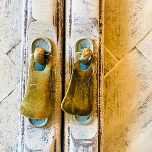 May include: Two antique brass cabinet door pulls with a worn, distressed finish. The pulls are attached to a white painted wooden cabinet door.