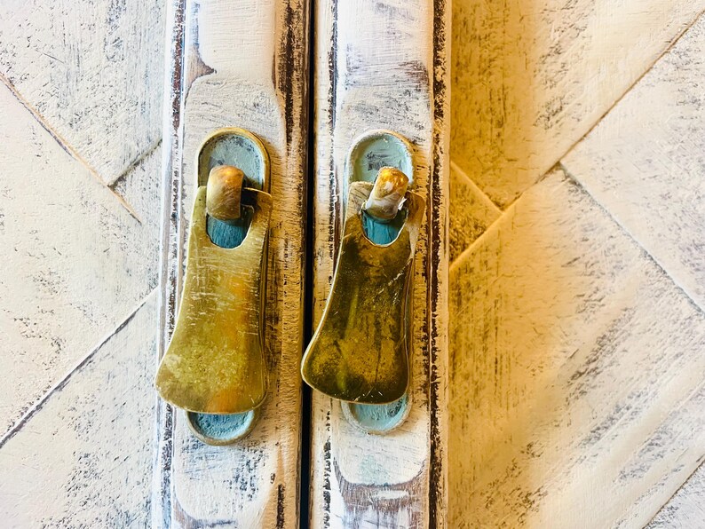 May include: Two antique brass cabinet door pulls with a worn, distressed finish. The pulls are attached to a white painted wooden cabinet door.