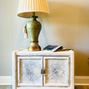 May include: A white painted wooden cabinet with two doors and brass hardware. The cabinet has a distressed finish and a diamond pattern on the doors. A green lamp with a white lampshade and a magazine are on top of the cabinet.