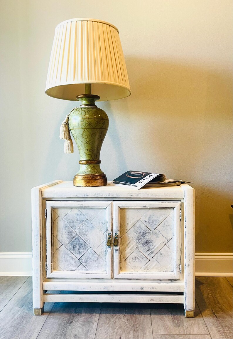 May include: A white painted wooden cabinet with two doors and brass hardware. The cabinet has a distressed finish and a diamond pattern on the doors. A green lamp with a white lampshade and a magazine are on top of the cabinet.