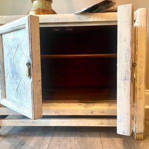May include: White painted wooden cabinet with a single door open, revealing a shelf inside. The cabinet has a distressed finish and gold-colored hardware.