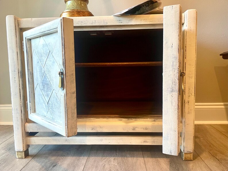 May include: White painted wooden cabinet with a single door open, revealing a shelf inside. The cabinet has a distressed finish and gold-colored hardware.