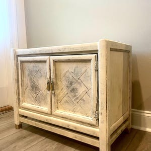 May include: A white wooden cabinet with two doors and a distressed finish. The doors have a diamond pattern and brass hardware. The cabinet is sitting on a light wood floor.
