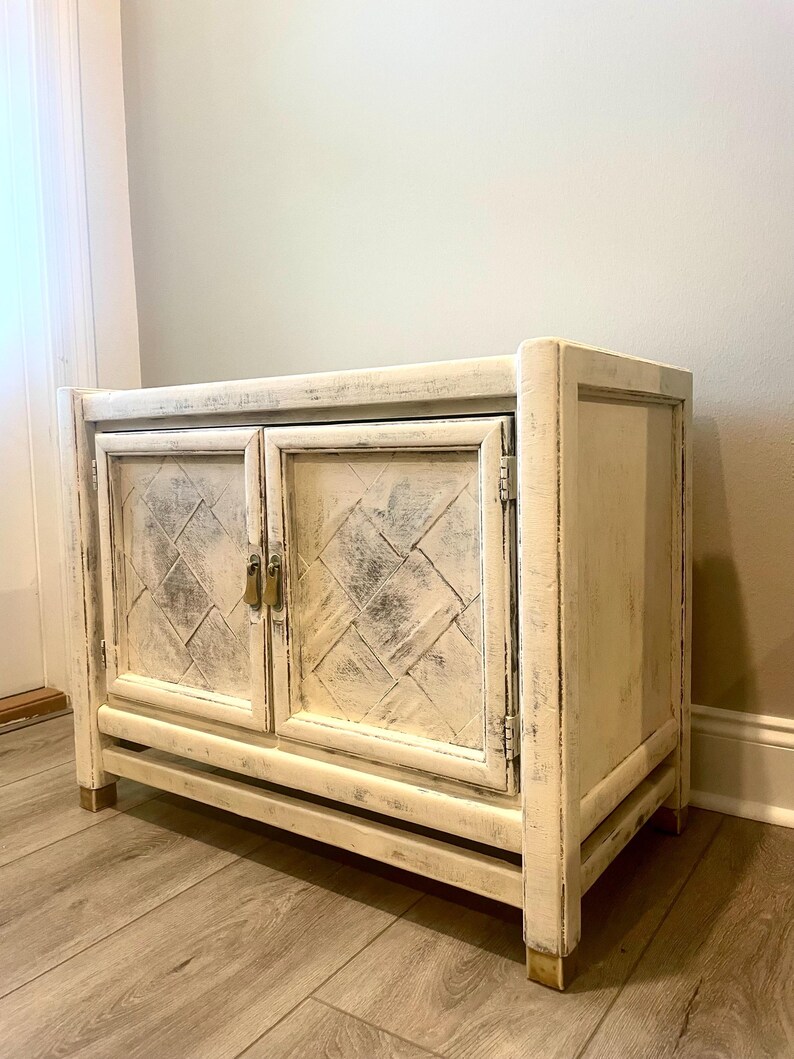May include: A white wooden cabinet with two doors and a distressed finish. The doors have a diamond pattern and brass hardware. The cabinet is sitting on a light wood floor.