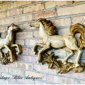 May include: Two vintage wall sculptures of horses in a running pose. The horses are a creamy beige color with gold-colored manes and tails. The sculptures are mounted on a brick wall. The text "Vintage. Blue Antiques" is at the bottom.