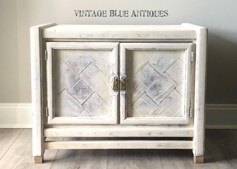 May include: White painted wooden cabinet with two doors and a distressed finish. The cabinet has a diamond pattern on the doors and brass hardware. "Vintage Blue Antiques" is printed on the wall behind the cabinet.