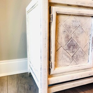 May include: White painted wooden cabinet with a distressed finish. The cabinet has a single door with a diamond pattern and brass feet.