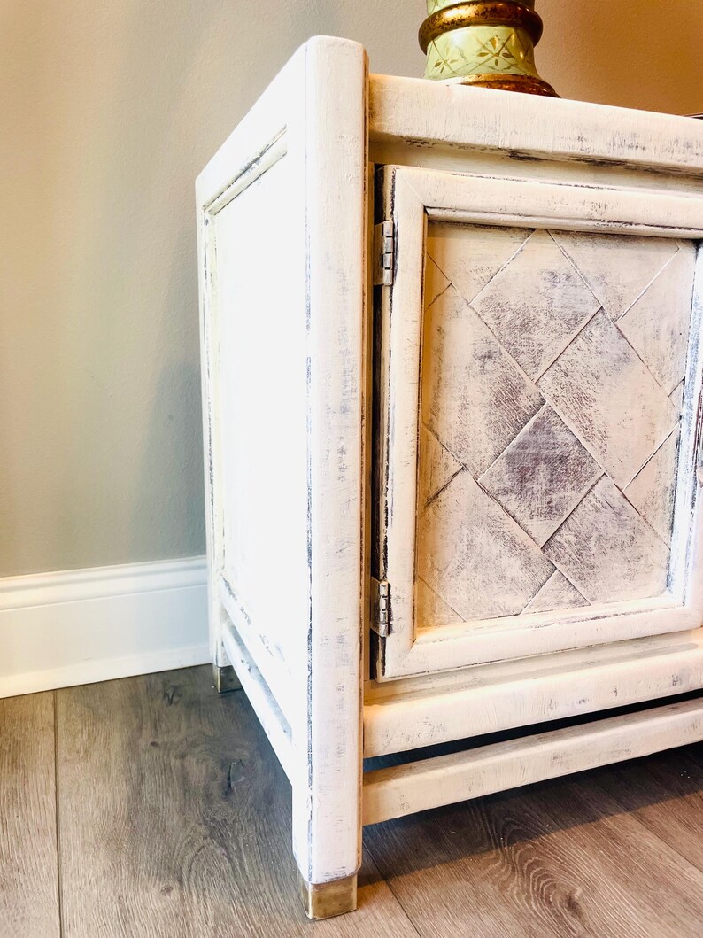 May include: White painted wooden cabinet with a distressed finish. The cabinet has a single door with a diamond pattern and brass feet.