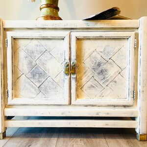 May include: White wooden cabinet with two doors and a diamond pattern on the front. The cabinet has brass hardware and sits on a wooden floor.