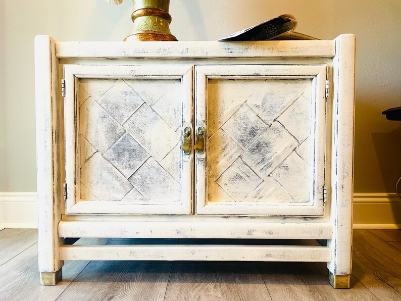 May include: White wooden cabinet with two doors and a diamond pattern on the front. The cabinet has brass hardware and sits on a wooden floor.