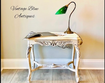 May include: White side table with ornate carvings and distressed paint. The table has a green lamp with a brass base on top. The table is sitting on a wooden floor.