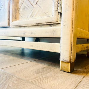 May include: A white distressed wooden cabinet with two doors and brass feet. The cabinet has a diamond pattern on the doors and is sitting on a wooden floor.