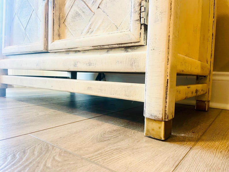 May include: A white distressed wooden cabinet with two doors and brass feet. The cabinet has a diamond pattern on the doors and is sitting on a wooden floor.