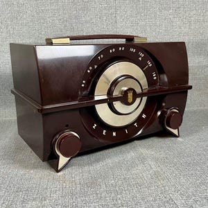 May include: A vintage brown Zenith radio with a silver dial and the brand name prominently displayed. The radio features a carrying handle and two control knobs. The dial displays numbers from 60 to 160. The radio is a classic piece of electronics.