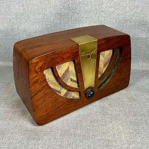 May include: A rectangular wooden radio with a polished dark brown finish. It features a central vertical brass plate engraved with the Zenith logo, a single black circular control knob, and two arched speaker grilles covered in a patterned fabric.