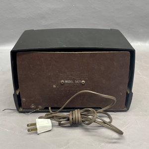 Op de afbeelding: Een zwarte vintage radio met een bruin luidsprekerrooster en een snoer met een stekker. De radio heeft een label met de tekst "MODEL 547A".