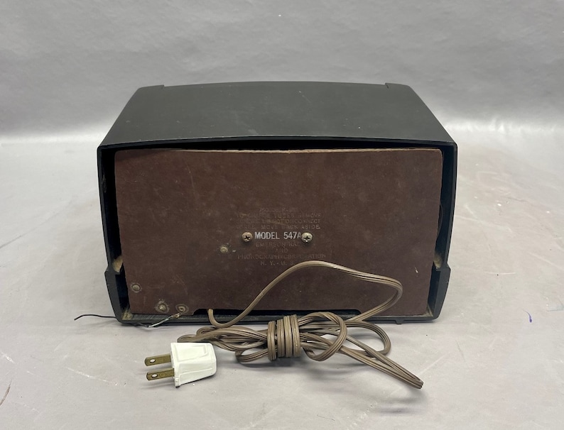 Op de afbeelding: Een zwarte vintage radio met een bruin luidsprekerrooster en een snoer met een stekker. De radio heeft een label met de tekst "MODEL 547A".
