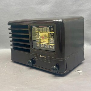 K&ouml;nnte beinhalten: Ein braunes Vintage-Radio von Emerson mit einem schwarzen Zifferblatt und einem verchromten Knopf. Das Zifferblatt hat eine Skala f&uuml;r AM-Radiofrequenzen mit Zahlen von 550 bis 1600 Kilohertz. Das Radio hat ein Lautsprechergitter an der Vorderseite und ein Holzkabinett.