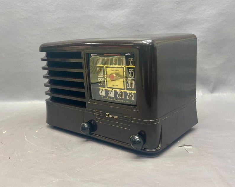 K&ouml;nnte beinhalten: Ein braunes Vintage-Radio von Emerson mit einem schwarzen Zifferblatt und einem verchromten Knopf. Das Zifferblatt hat eine Skala f&uuml;r AM-Radiofrequenzen mit Zahlen von 550 bis 1600 Kilohertz. Das Radio hat ein Lautsprechergitter an der Vorderseite und ein Holzkabinett.