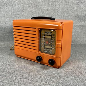 Puede incluir: Una radio vintage naranja con un asa y perillas negras. La radio tiene una cara rectangular con un dial que muestra n&uacute;meros y una aguja. El dise&ntilde;o incluye l&iacute;neas horizontales en el lado izquierdo.
