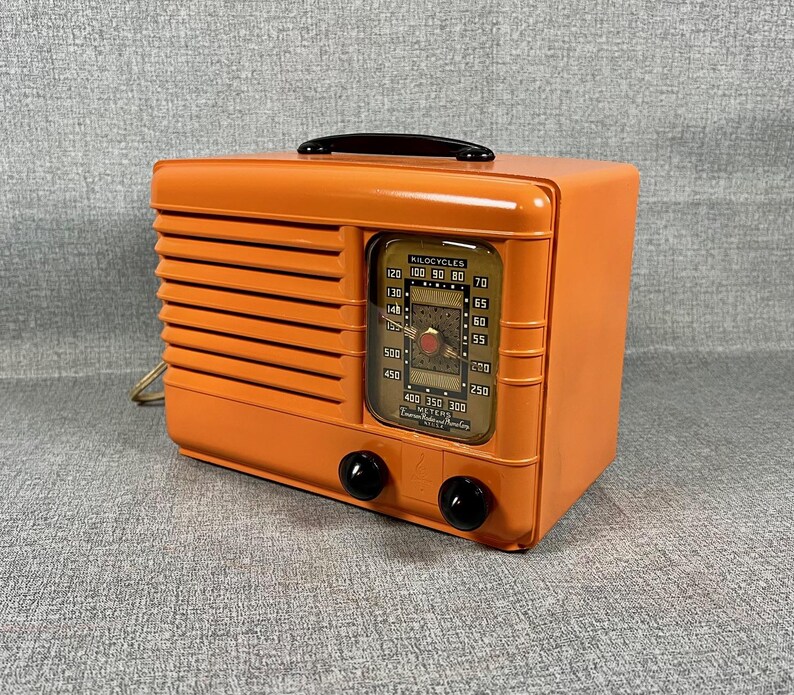 Puede incluir: Una radio vintage naranja con un asa y perillas negras. La radio tiene una cara rectangular con un dial que muestra n&uacute;meros y una aguja. El dise&ntilde;o incluye l&iacute;neas horizontales en el lado izquierdo.