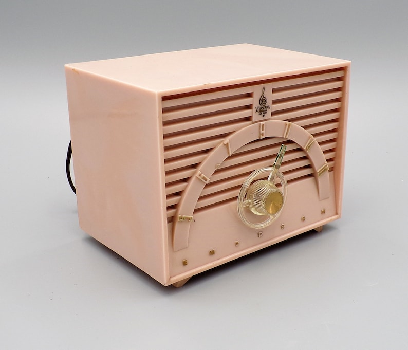 K&ouml;nnte beinhalten: Ein vintage-Radio von Emerson in rosa mit einem Drehknopf und einem "Emerson"-Label auf der Vorderseite. Das Radio hat ein Holzmaserungsmuster und ein "Duotone"-Logo mit einem Violinschl&uuml;ssel.