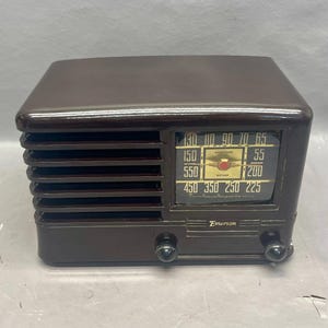 K&ouml;nnte beinhalten: Ein braunes Vintage-Radio von Emerson mit einem schwarzen Zifferblatt und verchromten Kn&ouml;pfen. Das Radio hat ein Lautsprechergitter mit vertikalen Lamellen und ein Zifferblatt mit Zahlen von 450 bis 70. Auf dem Zifferblatt steht auch "Automatic Radio Manufacturing Co. Inc."