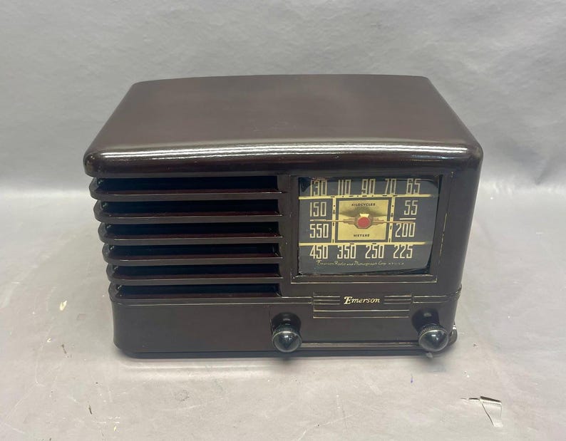 K&ouml;nnte beinhalten: Ein braunes Vintage-Radio von Emerson mit einem schwarzen Zifferblatt und verchromten Kn&ouml;pfen. Das Radio hat ein Lautsprechergitter mit vertikalen Lamellen und ein Zifferblatt mit Zahlen von 450 bis 70. Auf dem Zifferblatt steht auch "Automatic Radio Manufacturing Co. Inc."