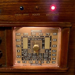 May include: Close-up of an antique radio dial with a copper-colored face and a wooden frame. The dial displays numbers, country names, and the words "Short Wave," "Police," and "Broadcast." A red light is illuminated.