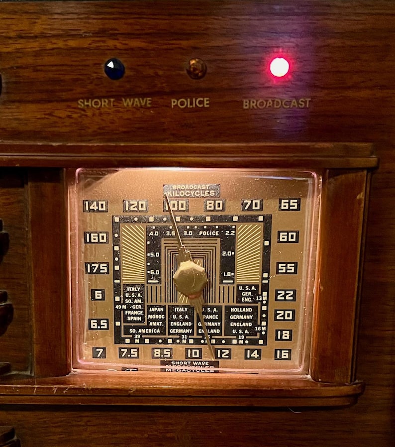 May include: Close-up of an antique radio dial with a copper-colored face and a wooden frame. The dial displays numbers, country names, and the words "Short Wave," "Police," and "Broadcast." A red light is illuminated.