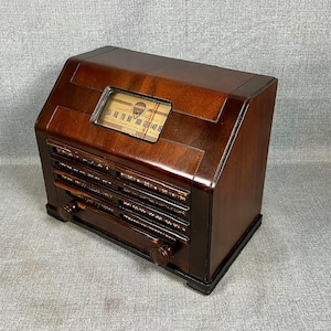 May include: A dark brown wooden radio with a slanted front panel, featuring a rectangular glass dial with frequency numbers ranging from 55 to 160, two circular control knobs at the bottom, and horizontal slats covering the speaker area.