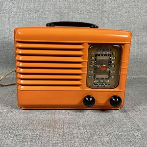 Puede incluir: Una radio naranja vintage con un asa negra y dos perillas negras. La radio tiene un dial con n&uacute;meros y una aguja. La parte frontal de la radio tiene l&iacute;neas horizontales y un cable de alimentaci&oacute;n.