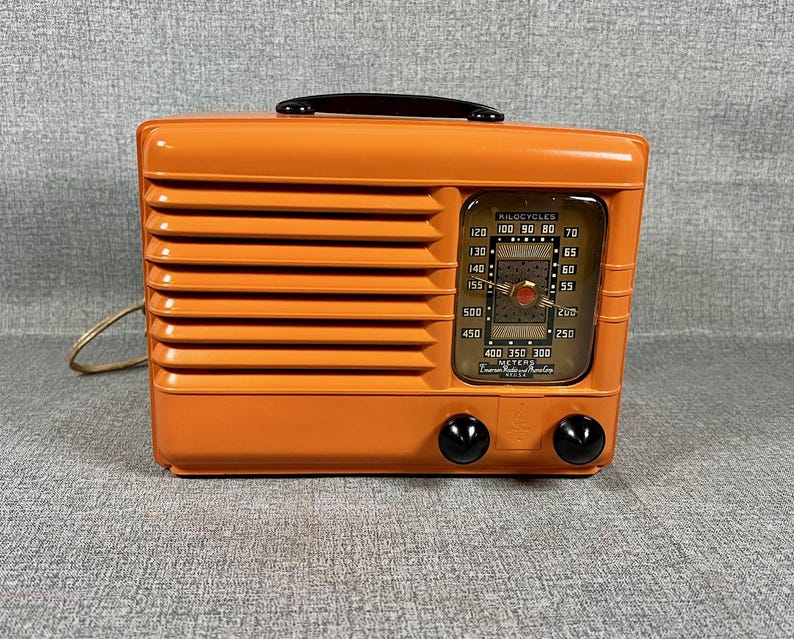 Puede incluir: Una radio naranja vintage con un asa negra y dos perillas negras. La radio tiene un dial con n&uacute;meros y una aguja. La parte frontal de la radio tiene l&iacute;neas horizontales y un cable de alimentaci&oacute;n.