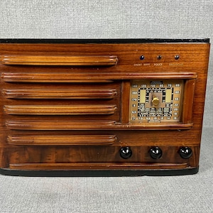 May include: Vintage wooden radio with a dark finish. The radio features a rectangular design with horizontal grooves on the front and a dial with numbers and a tuning knob. The text "SHORT WAVE POLICE" is visible above the dial. Three black knobs are at the bottom.