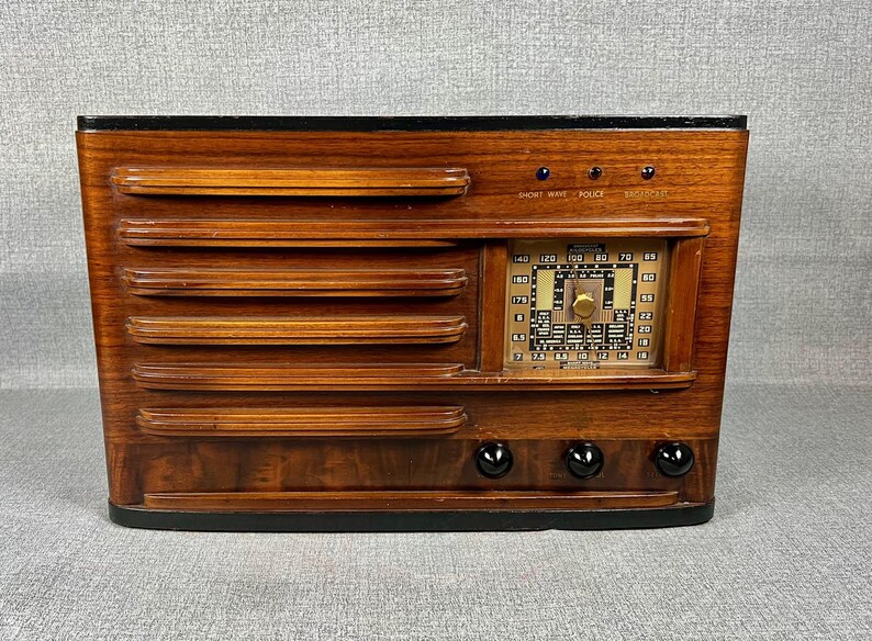 May include: Vintage wooden radio with a dark finish. The radio features a rectangular design with horizontal grooves on the front and a dial with numbers and a tuning knob. The text "SHORT WAVE POLICE" is visible above the dial. Three black knobs are at the bottom.