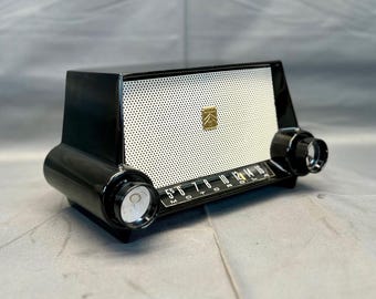 May include: Vintage black and white Motorola radio with a perforated speaker grill and gold emblem. The radio features two large black knobs and a frequency dial. The dial reads "Motorola" and numbers from 55 to 16.