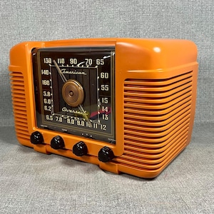 May include: A bright orange vintage radio with a rectangular black dial featuring the text American Overseas. The radio has horizontal side vents, four black control knobs along the bottom, and a central circular dial with a silver needle indicator.