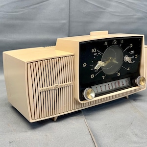 May include: A vintage cream-colored General Electric clock radio with a black clock face and a silver tuning knob. The radio has a wood-grain design and a speaker grille.