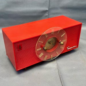 May include: A red vintage Emerson clock radio with a clear plastic face and gold-colored hands. The radio has a textured surface and the word "Emerson" is printed on the front.