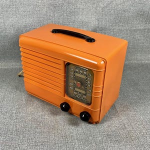 Puede incluir: Una radio vintage naranja con un asa negra y dos perillas negras. La radio tiene forma rectangular con una rejilla de altavoz en el lateral y un dial con n&uacute;meros y marcas. La radio es de estilo retro.