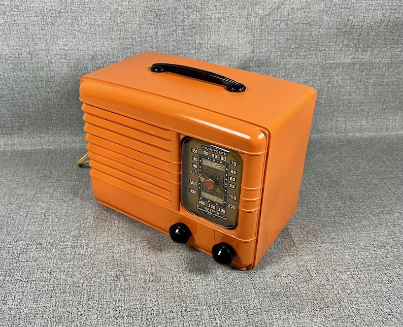 Puede incluir: Una radio vintage naranja con un asa negra y dos perillas negras. La radio tiene forma rectangular con una rejilla de altavoz en el lateral y un dial con n&uacute;meros y marcas. La radio es de estilo retro.