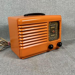 Puede incluir: Radio vintage naranja con asa negra y dos perillas negras. La radio tiene una rejilla de altavoz en la parte delantera y un dial con n&uacute;meros y una aguja. Un cable de alimentaci&oacute;n se extiende desde el lateral.