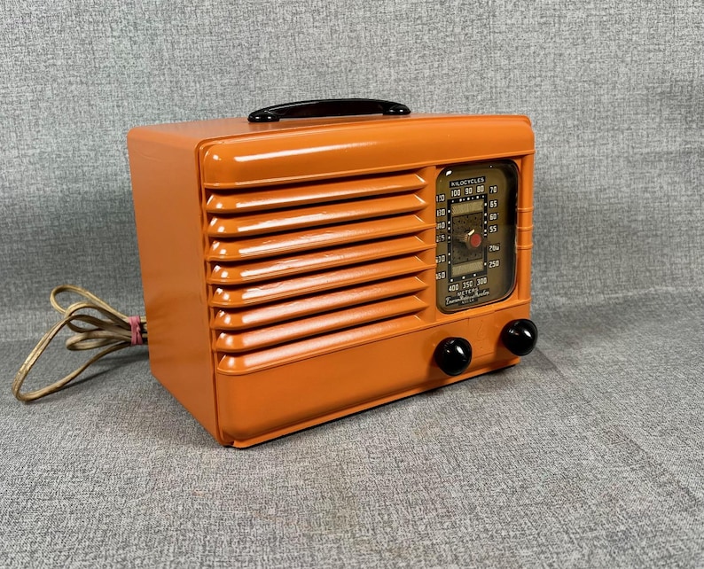 Puede incluir: Radio vintage naranja con asa negra y dos perillas negras. La radio tiene una rejilla de altavoz en la parte delantera y un dial con n&uacute;meros y una aguja. Un cable de alimentaci&oacute;n se extiende desde el lateral.
